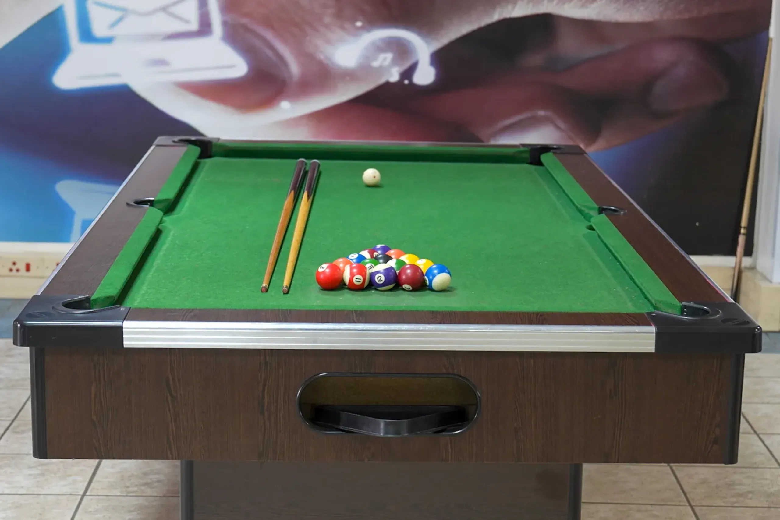Pool table with green felt, two cue sticks resting on the table, and a cluster of billiard balls near the far end.