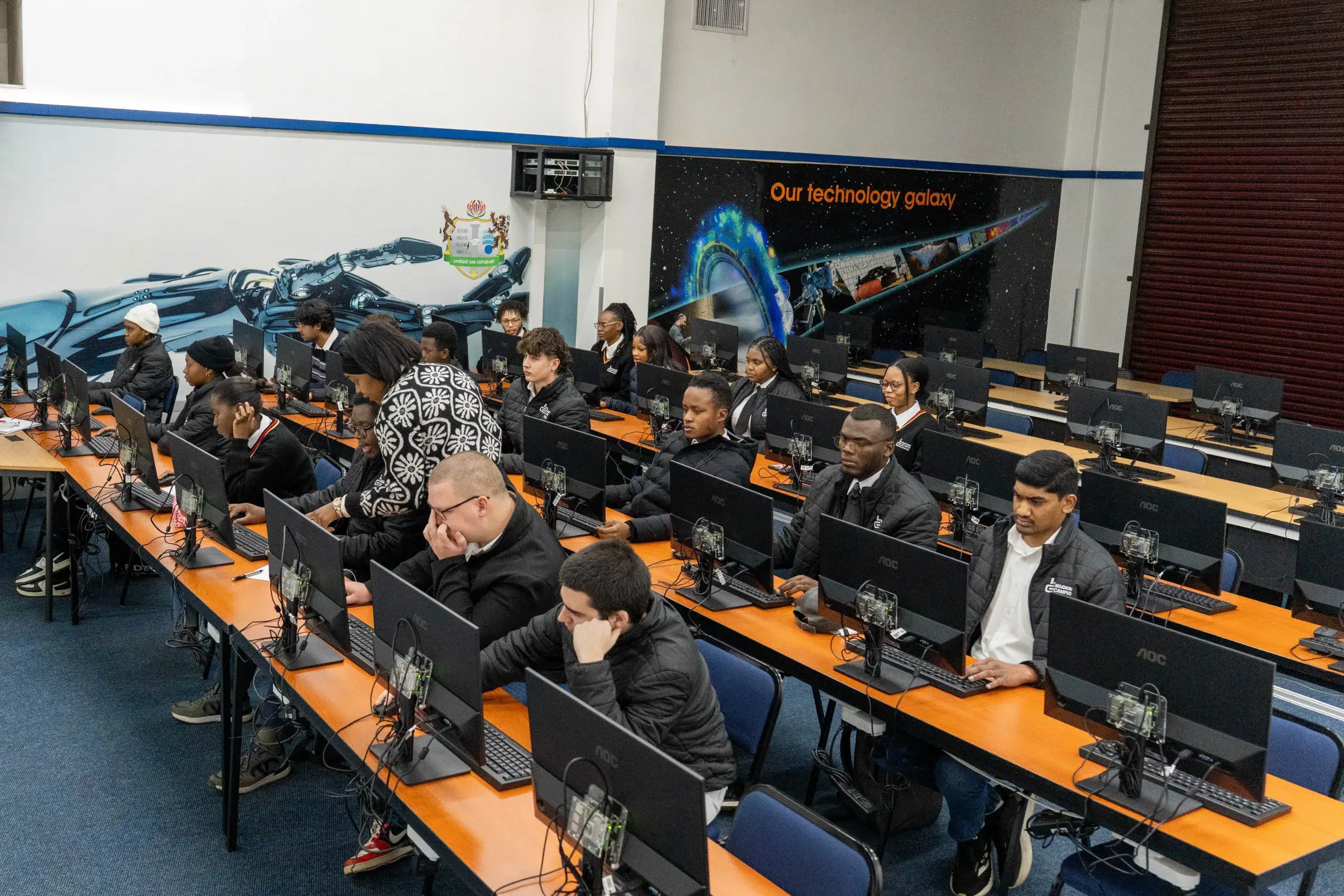 A computer lab full of students at desks with monitors and keyboards, several wearing jackets, focused on screens.