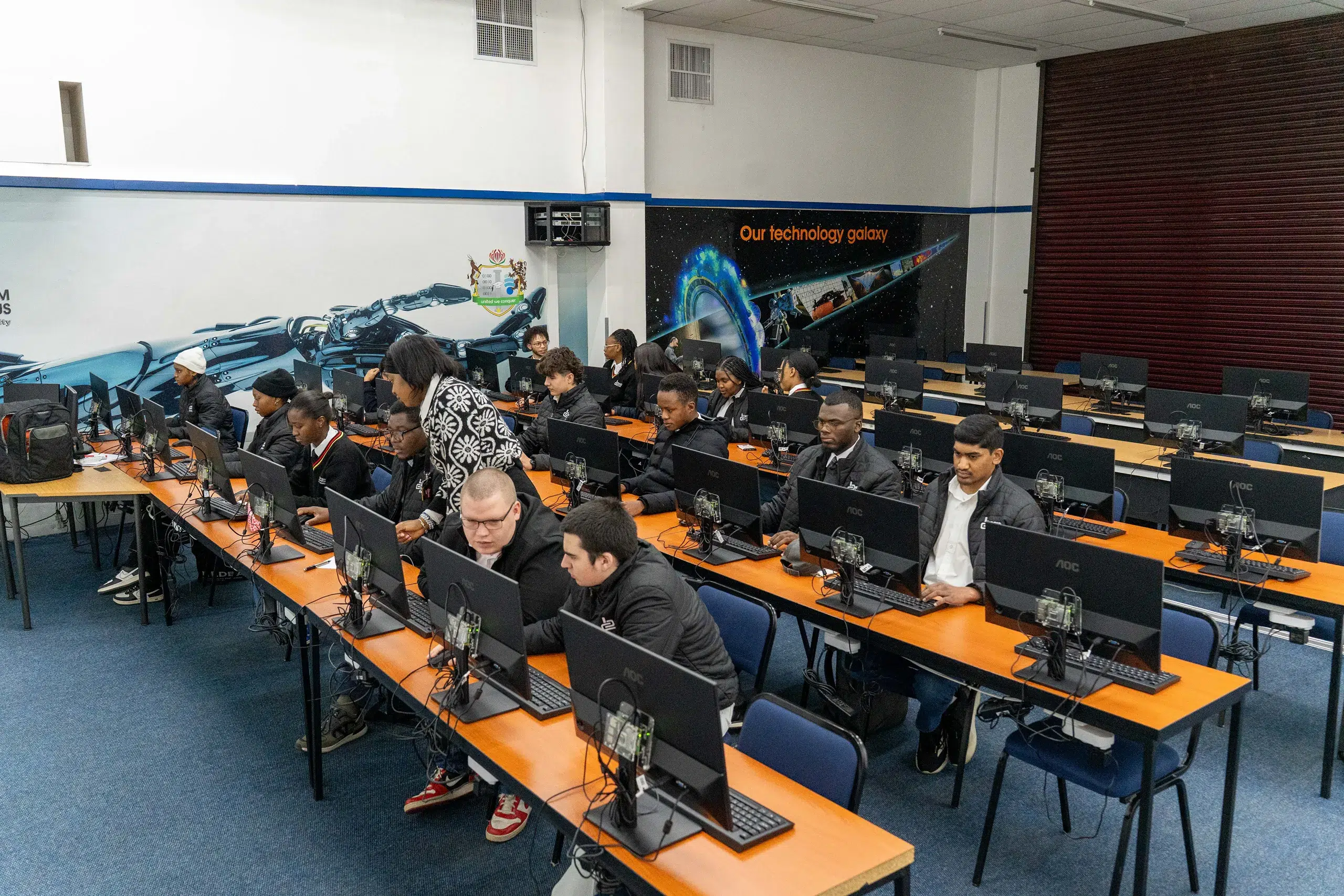 Students working on desktop computers in a computer lab with orange desks and black monitors.