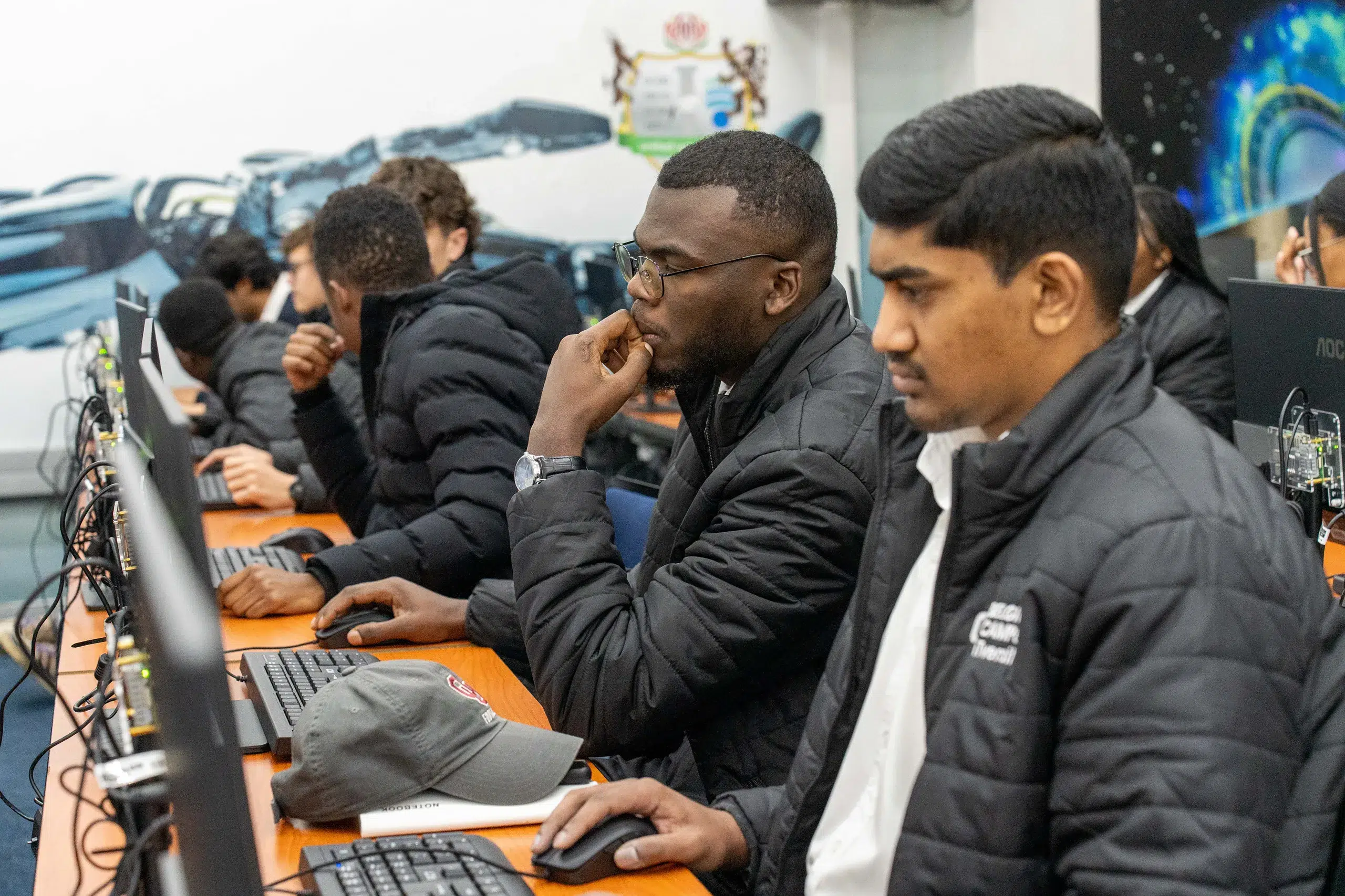 Group of students in a computer lab working at desktop PCs, with a central man in glasses touching his lips while seated at a workstation.