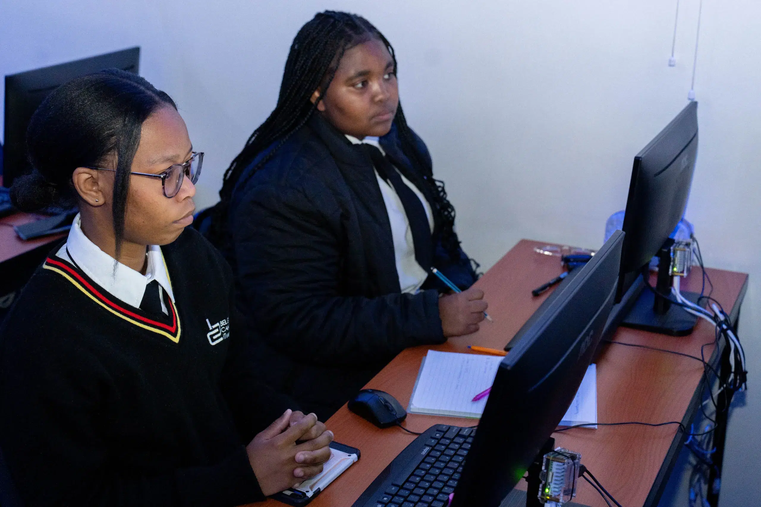 Two students in school uniforms sit at a desk with desktop computers, taking notes while looking at screens in a computer lab.