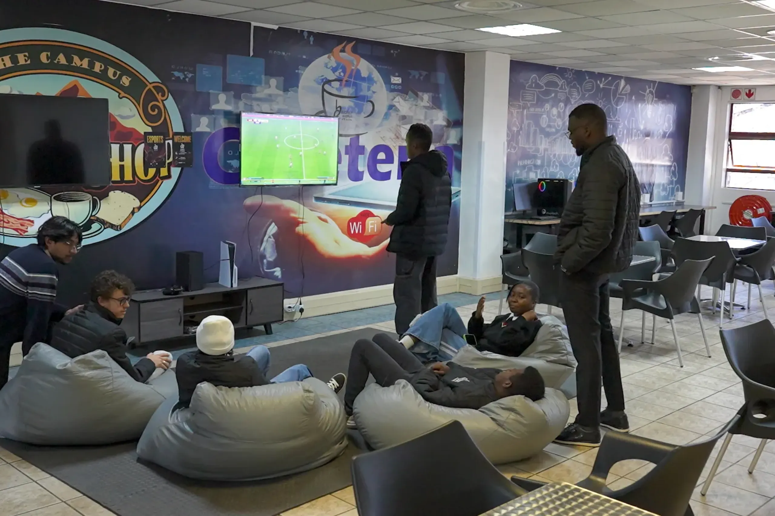 Students in a campus lounge relax on bean bags while a soccer video game plays on a wall-mounted screen wisely.