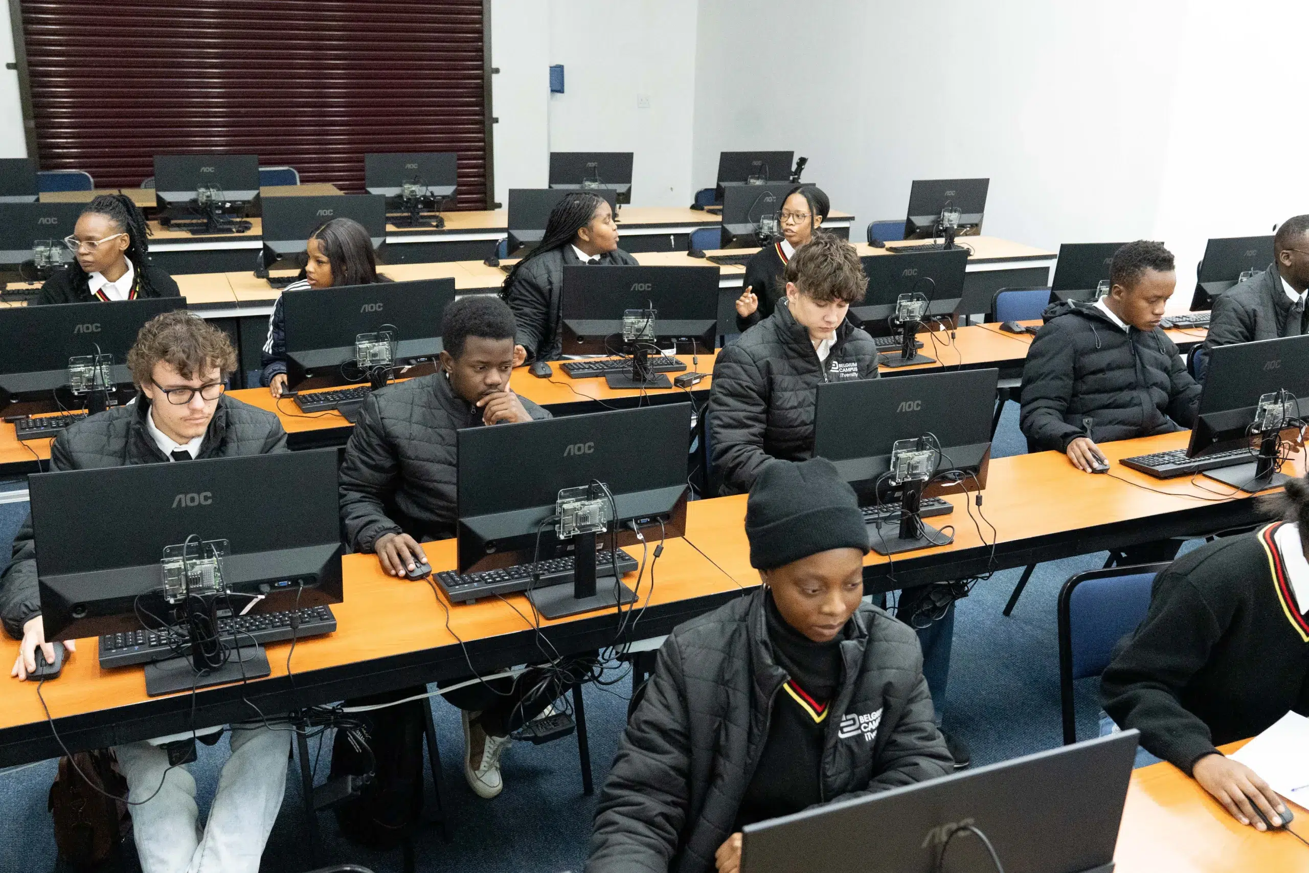 Students sit at computer lab desks using desktop PCs in a classroom, focused on screens.