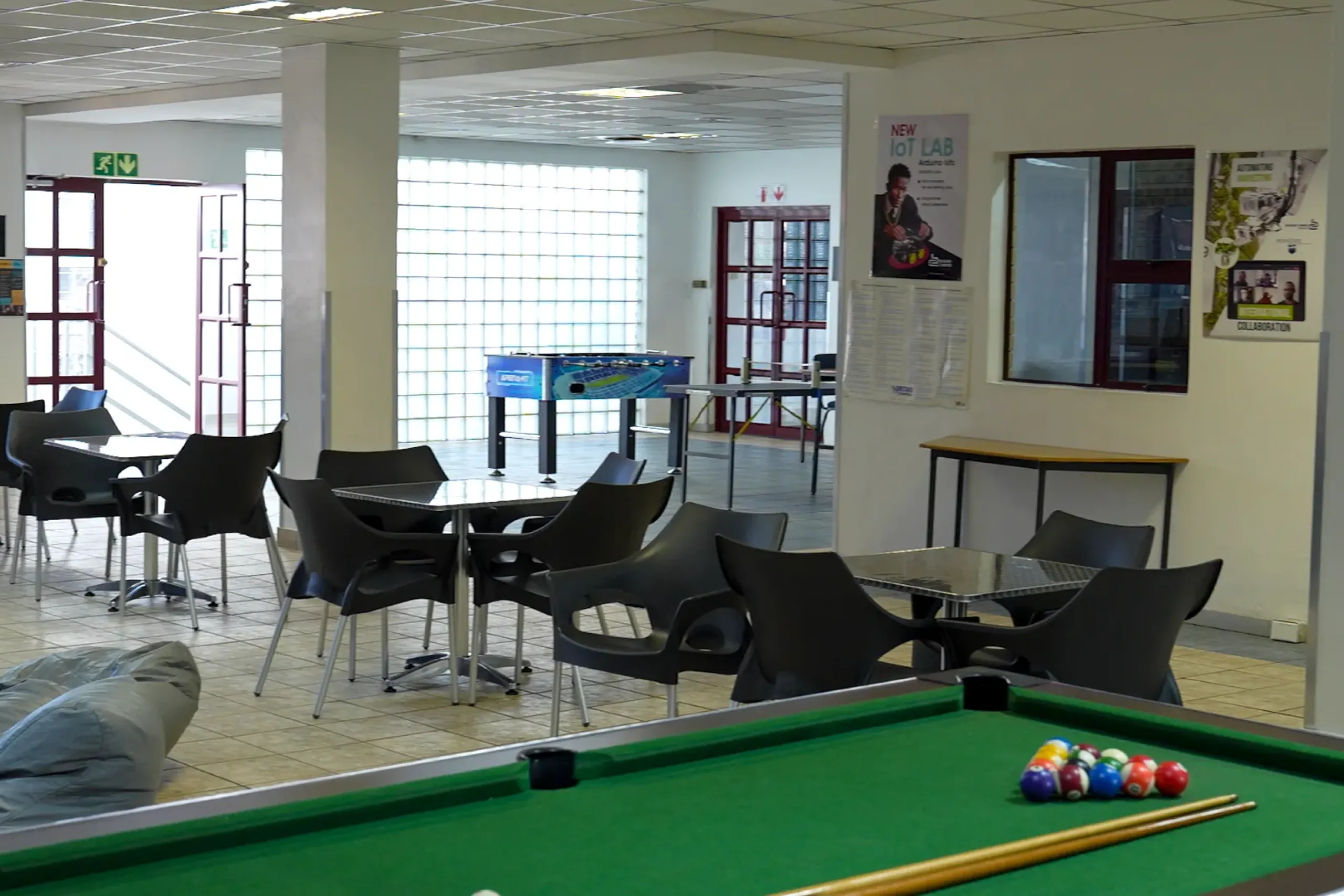 Indoor recreational area with a green billiards table in the foreground and black chairs around tables; posters on the wall and glass block window in the background.