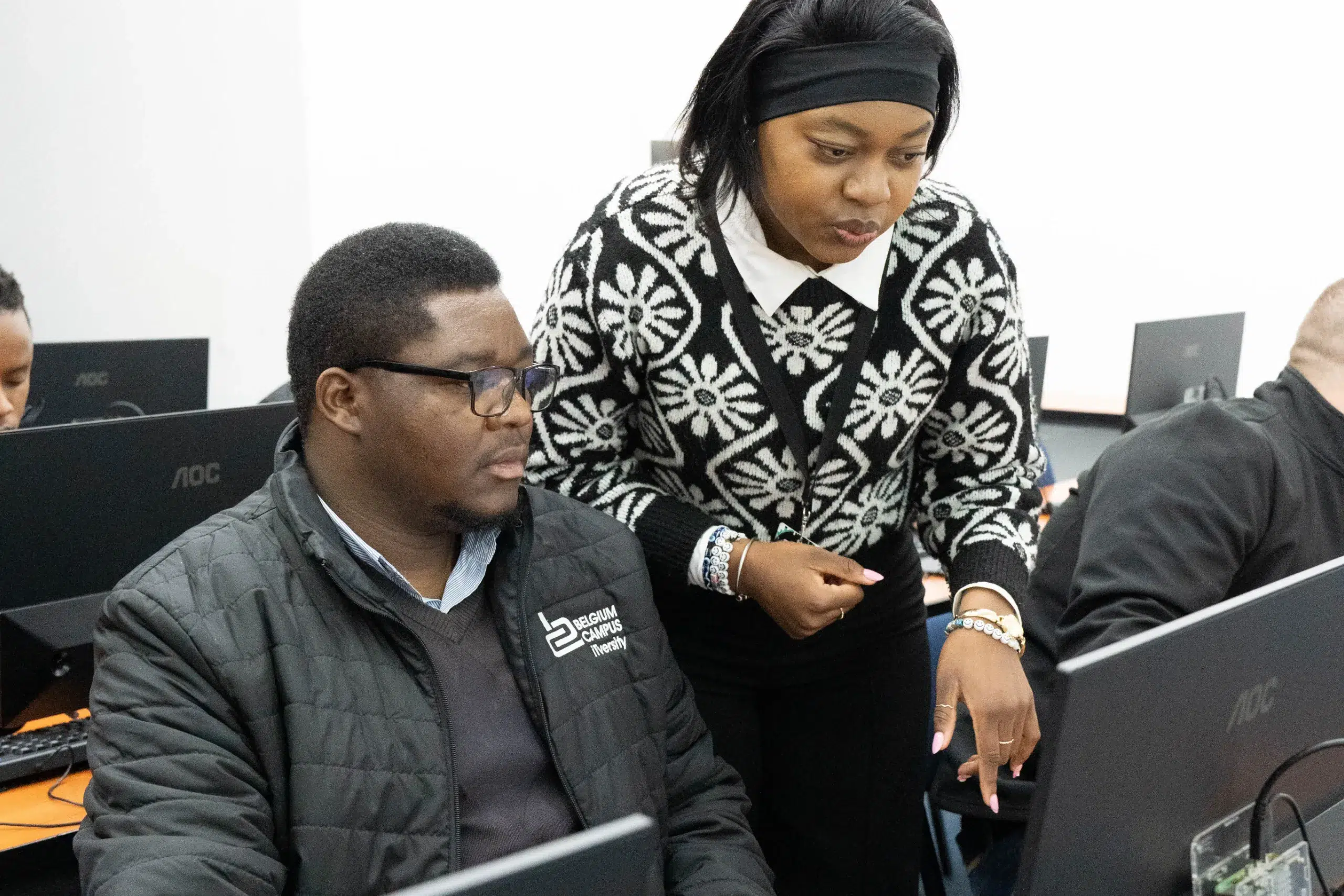 Man seated at a computer in a classroom, with a woman standing beside him leaning over to help him. She wears a black headband and a black-and-white patterned sweater.",