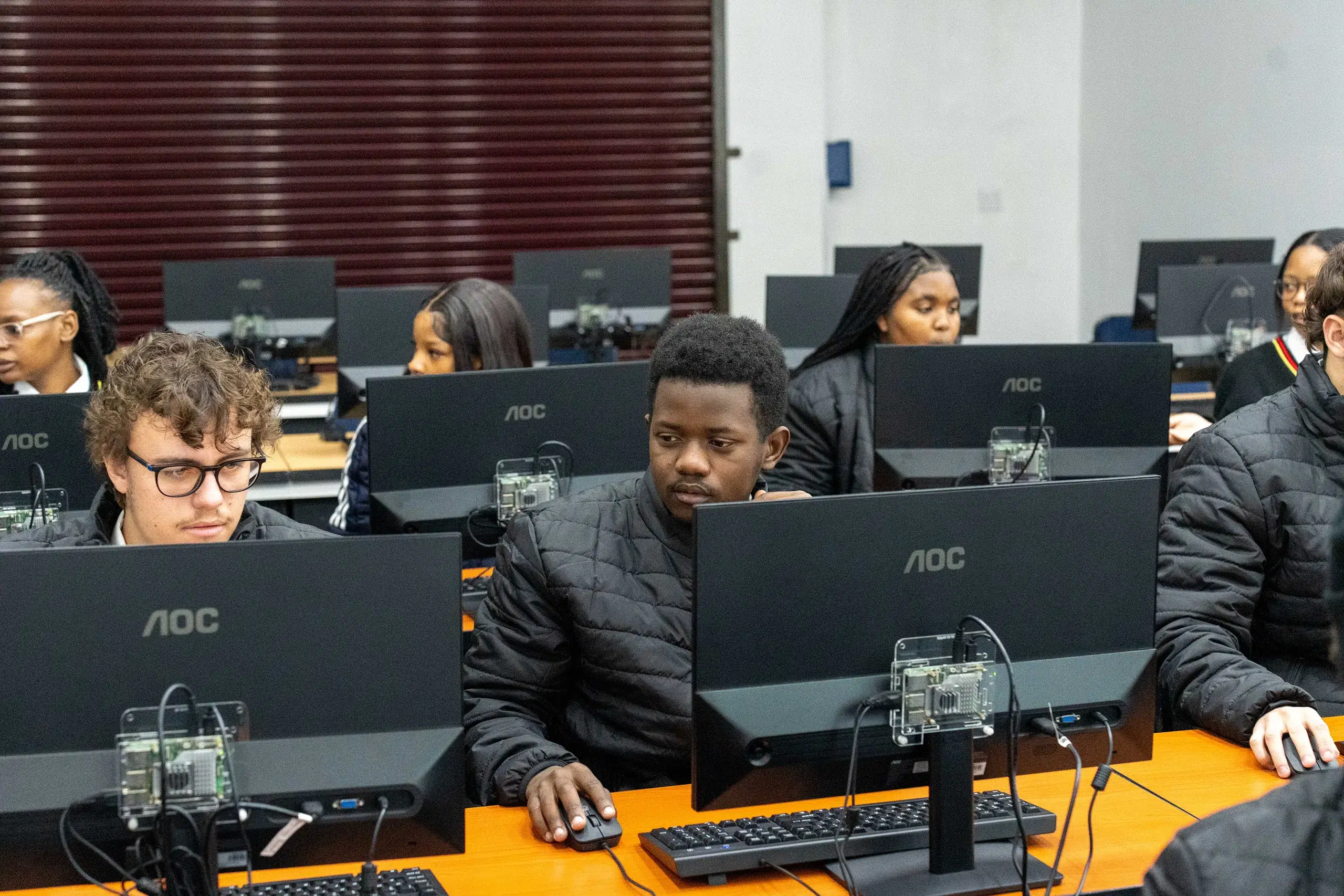 Students in a computer lab focused on desktop workstations at a long orange table.