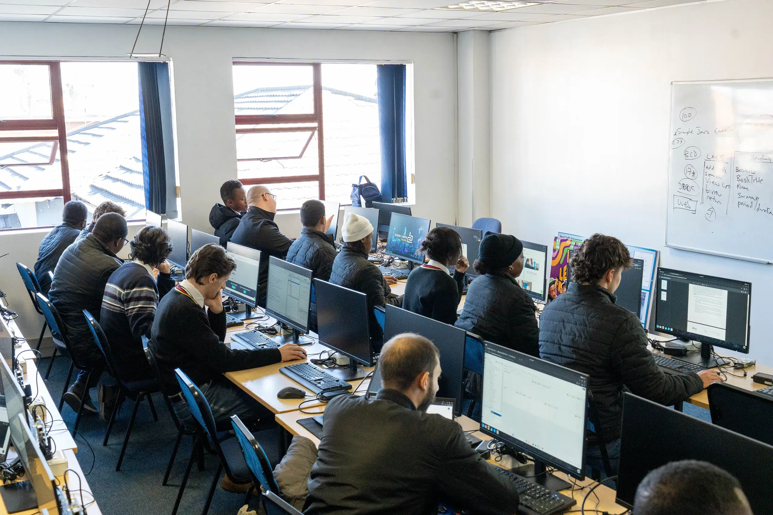 Students in a computer lab seated at long desks using desktop computers, with a whiteboard on the wall.