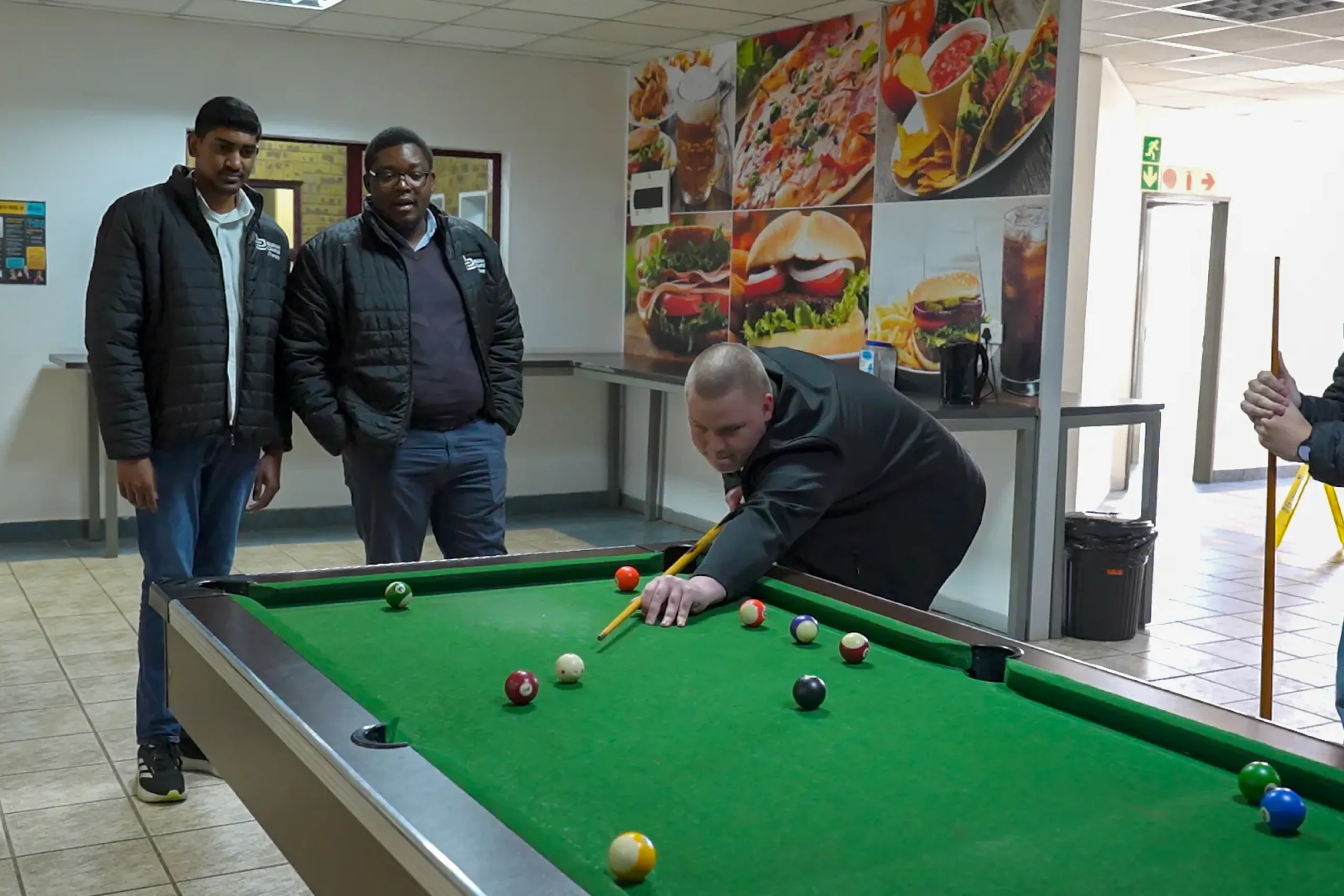 Man bends over a billiards table to take a shot as two men stand nearby, watching, in a room with large food posters on the wall.