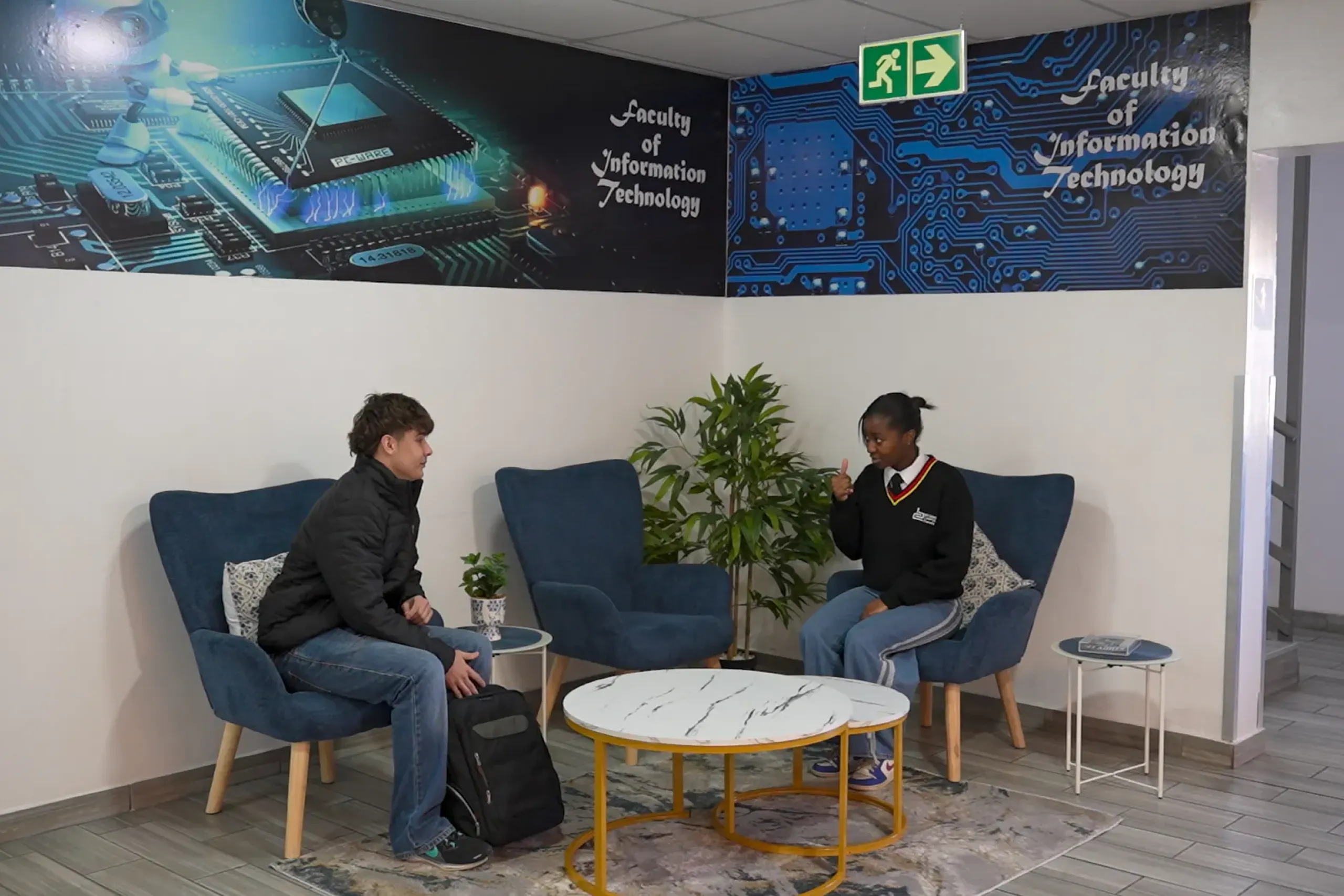Two students sit on blue chairs in a tech-themed lounge, chatting near a round marble-top table and plants—wall art reads 'Faculty of Information Technology'.