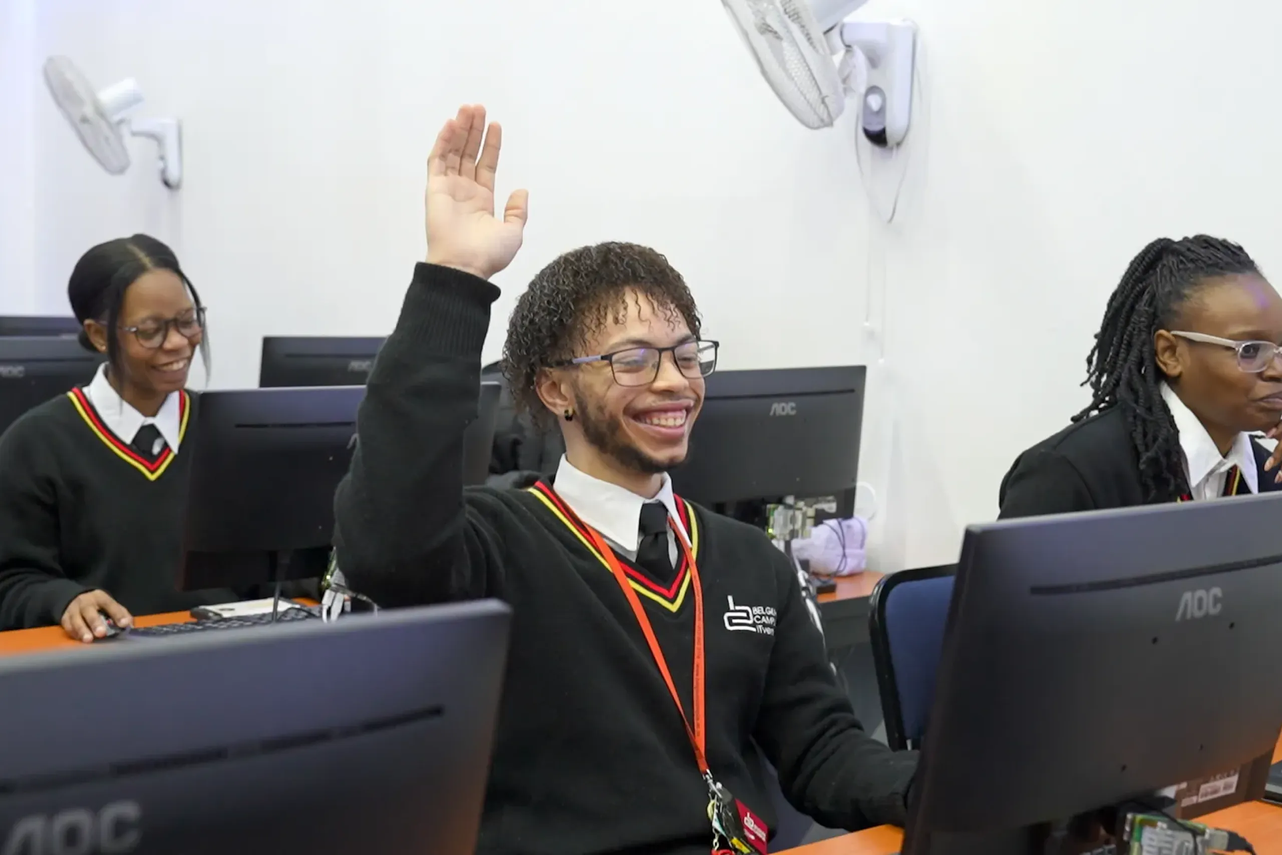 Student in a black uniform raises his hand at a computer desk in a bright classroom, others working nearby.