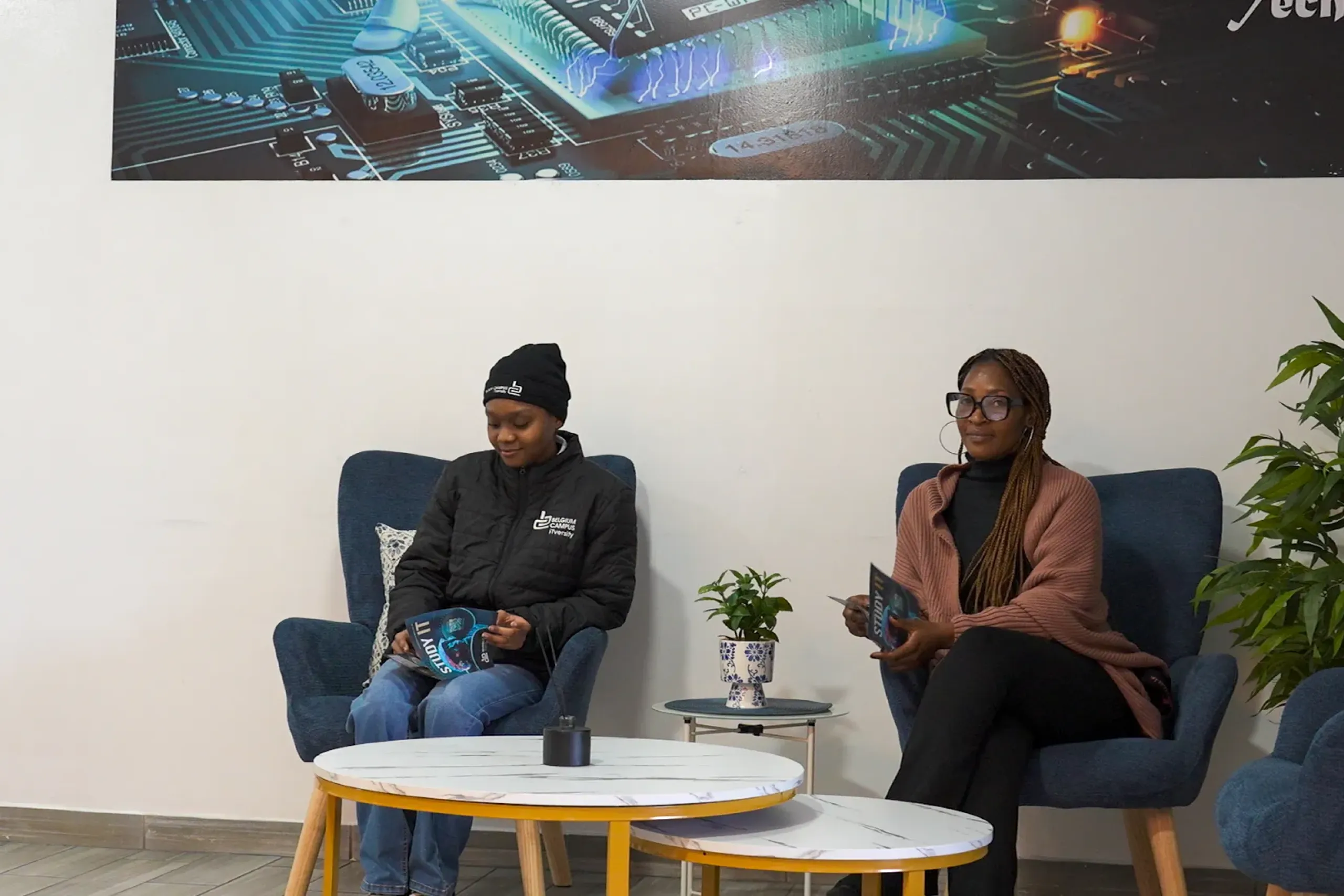 Two people sit in blue lounge chairs reading magazines in a modern waiting area, with a small round table and a potted plant nearby.
