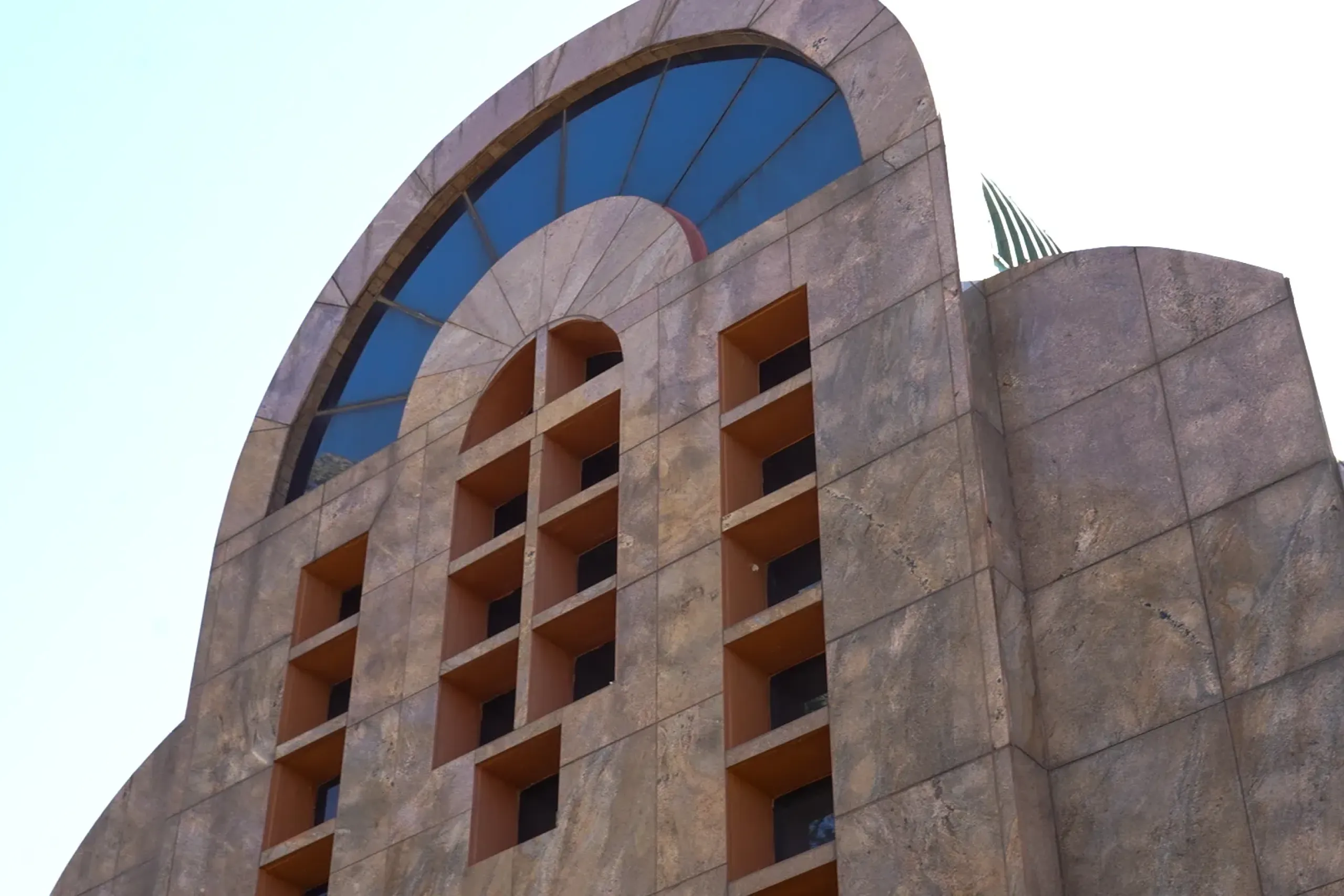 Facade of a modern beige stone building with a large arched blue window and orange rectangular recesses.