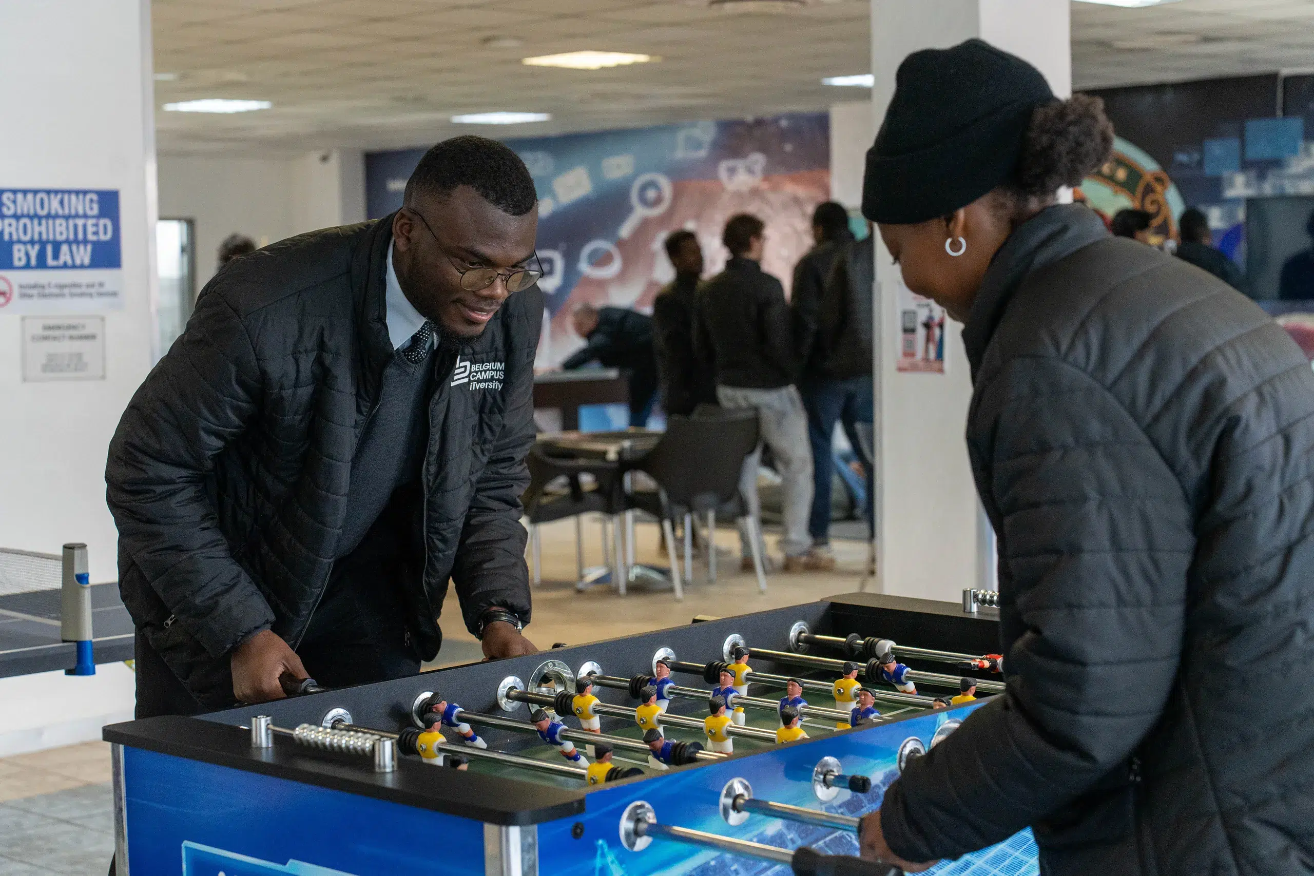 Two people play foosball indoors, leaning over a blue table with yellow and blue players, as others watch in the background.