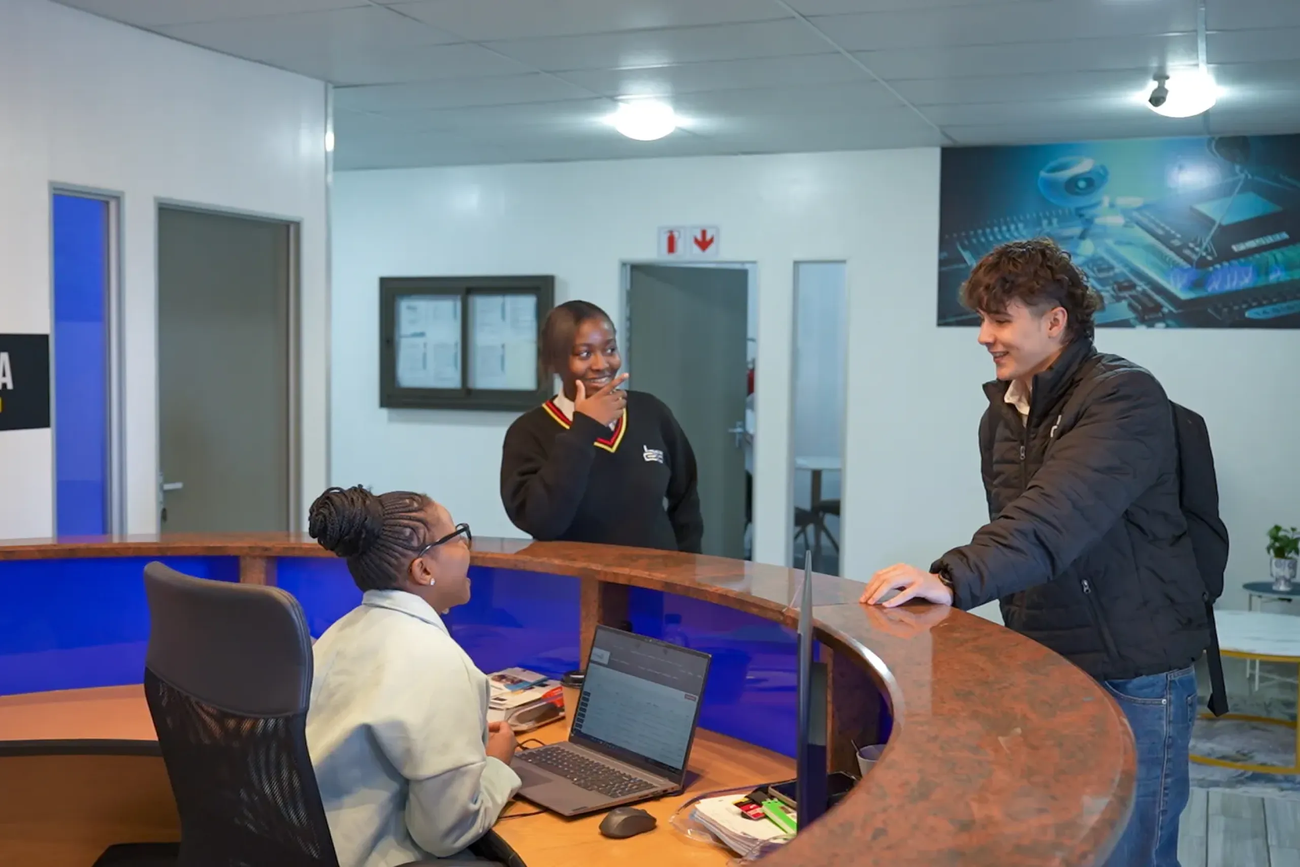 Student seated with a laptop at a curved wooden desk as two visitors chat at a bright reception in a modern lobby.