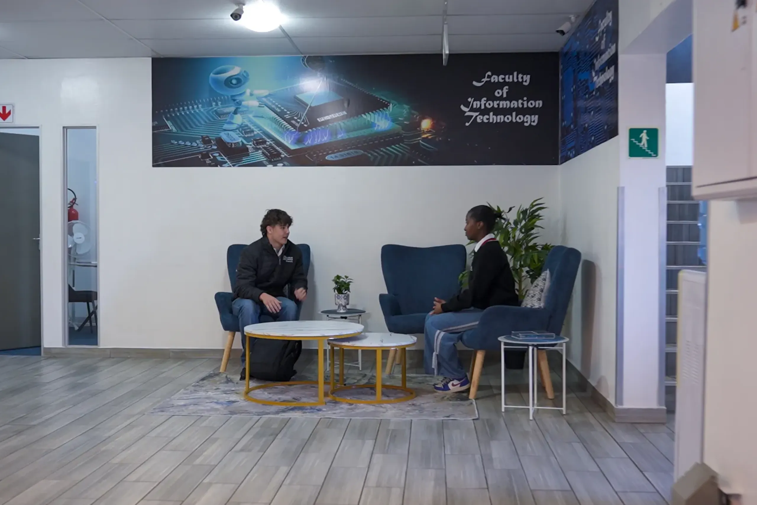 Two people sit in blue lounge chairs across from each other in a tech-themed lobby with round yellow tables and a plant, chatting casually.