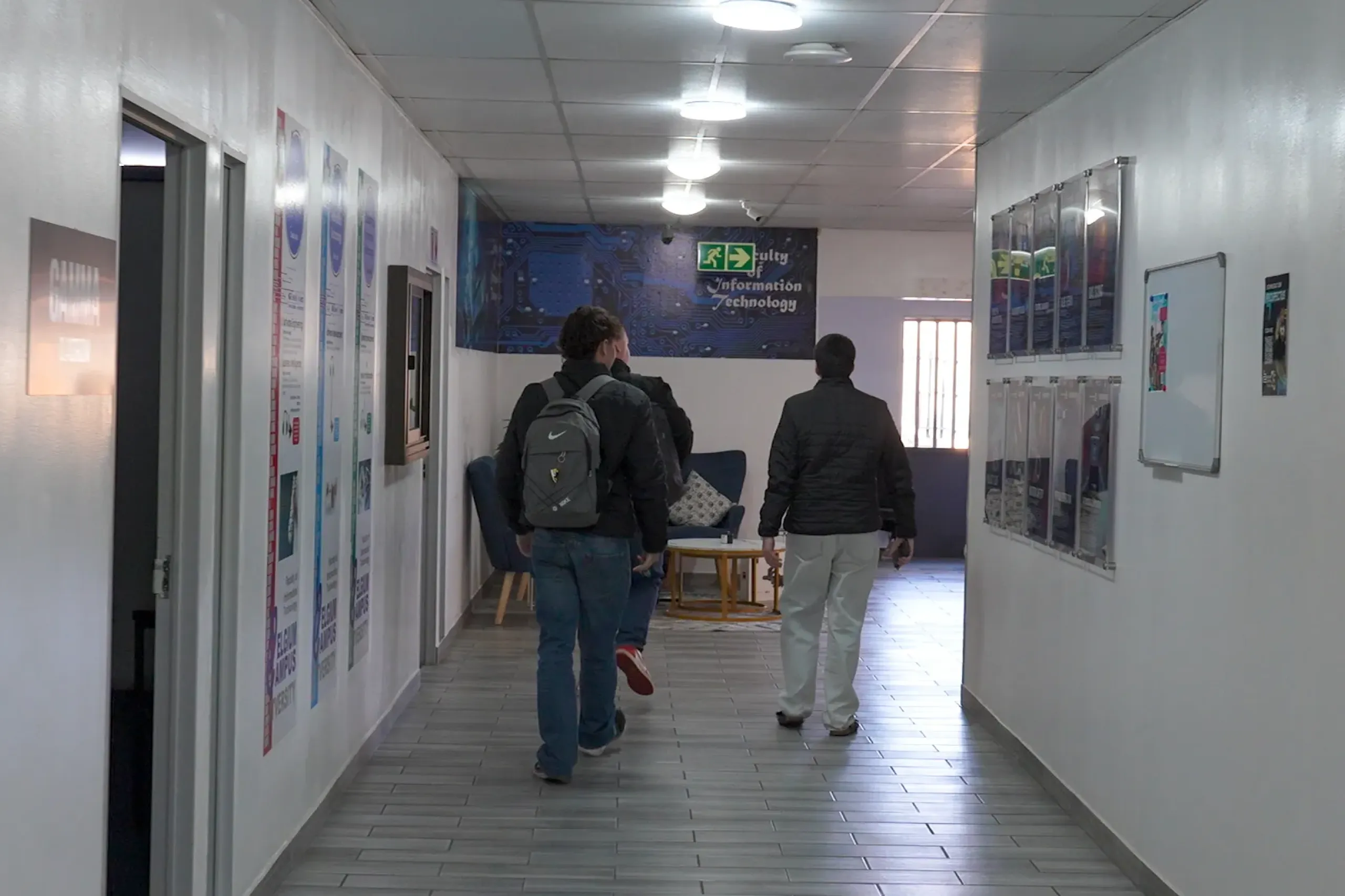 Three students wearing jackets with backpacks walk down a well-lit hallway lined with posters on white walls, toward a blue mural at the far end.