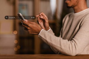 Person seated at a table using a smartphone, with a transparent search bar overlay displayed in the foreground. The search bar contains a search icon, the placeholder text "Search...", and microphone and camera icons, suggesting voice and visual search features. The background is softly blurred with warm indoor lighting.