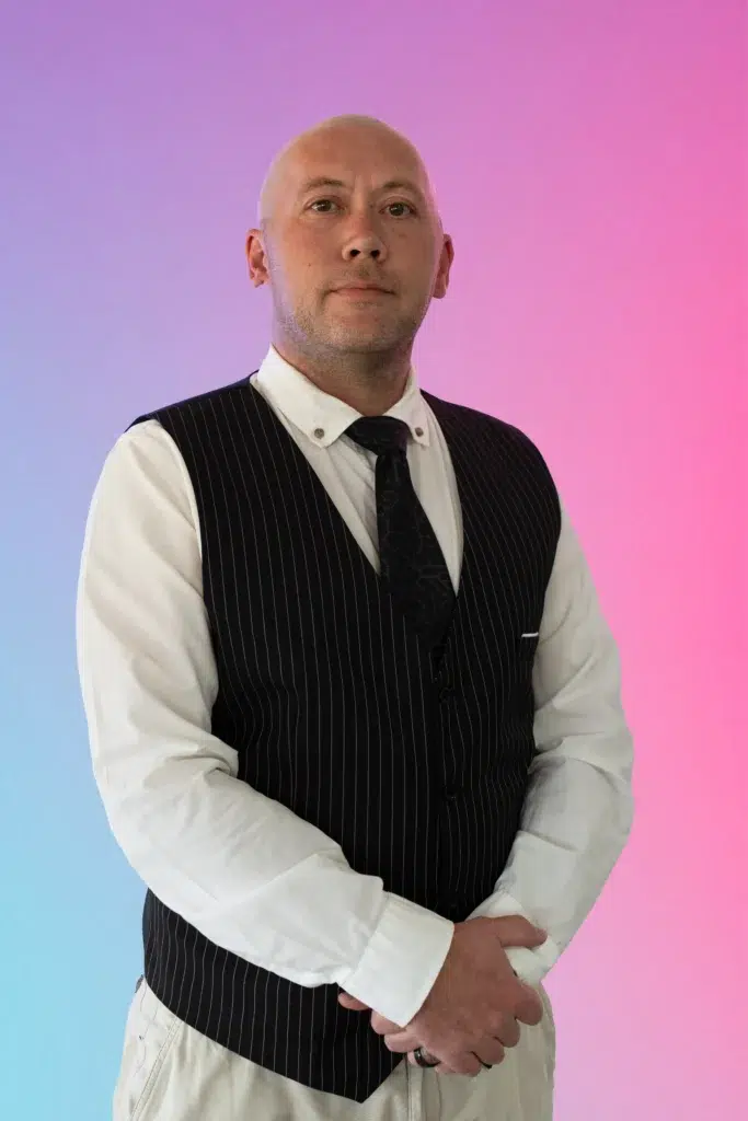 Portrait of a bald man in a white dress shirt, black pinstripe vest, and tie, standing with hands loosely clasped against a pink-blue gradient background.
