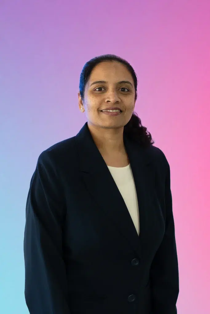 Portrait of a woman with dark hair tied back, wearing a navy blazer and white top, smiling against a pink–blue gradient backdrop.