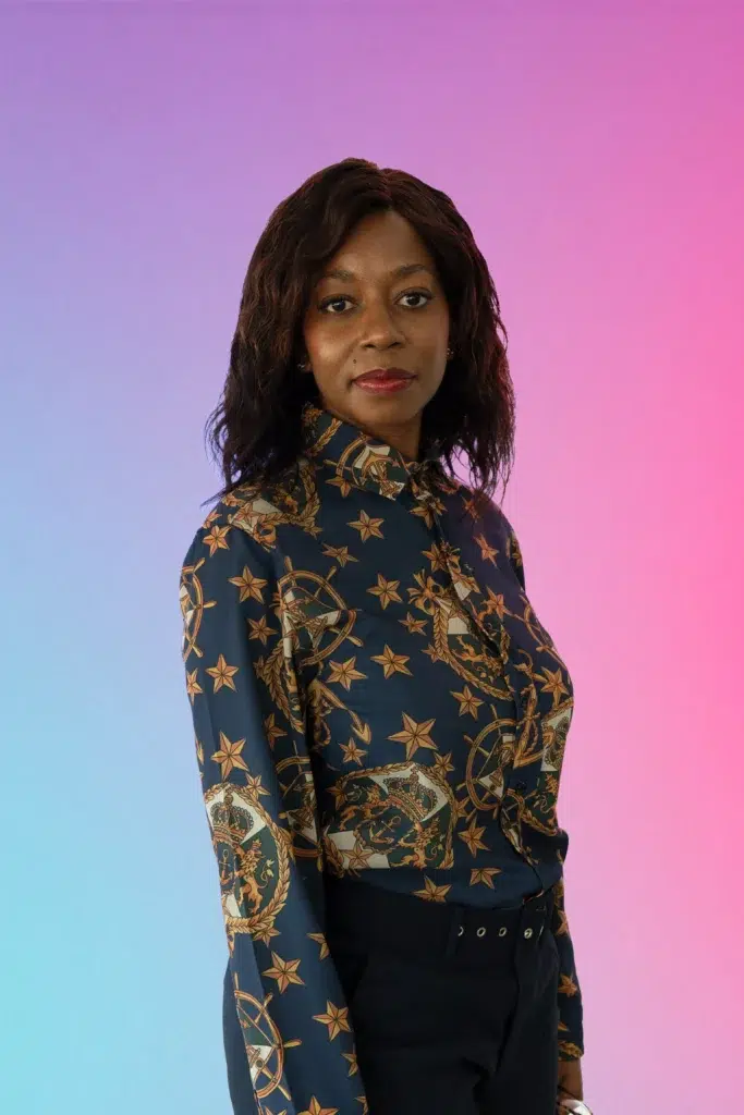 Portrait of a woman with wavy dark hair wearing a navy patterned blouse, standing against a pink‑to‑blue gradient background