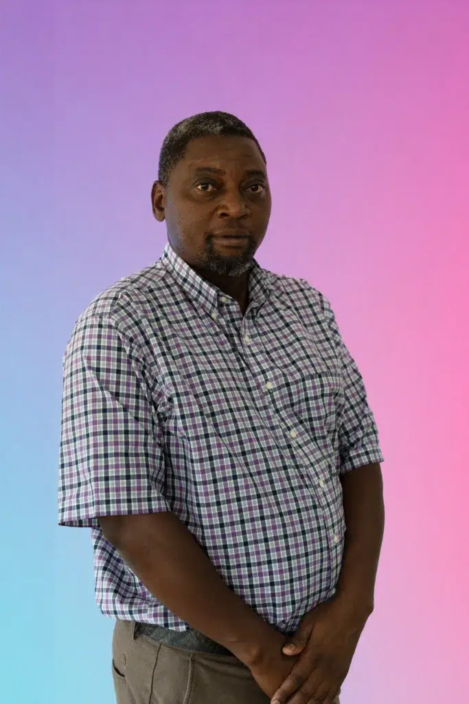 Portrait of a man in a purple plaid shirt standing against a pink-to-blue gradient background.