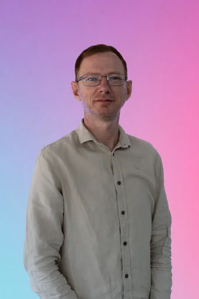 Portrait of a man wearing glasses and a light beige button-down shirt against a pink-to-blue gradient background, looking at the camera.