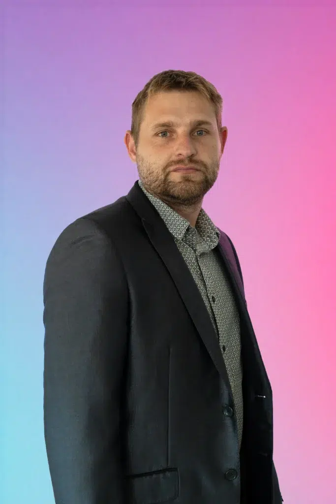 Portrait of a man in a dark blazer and patterned shirt against a pink-to-blue gradient background.