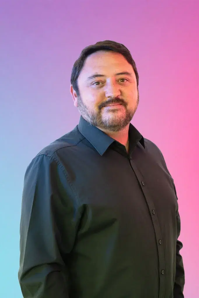 Portrait of a man with dark hair and a beard wearing a dark shirt, posed against a blue-to-pink gradient background.