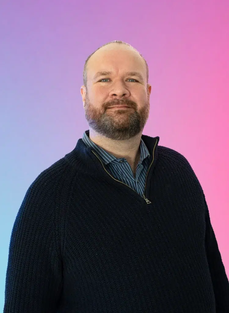Portrait of a man with short hair and beard wearing a dark knit sweater over a striped blue shirt, against a pink–purple gradient background.