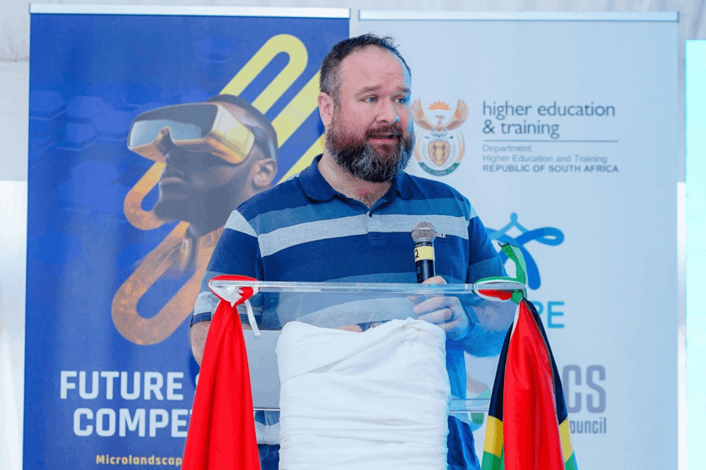 A speaker stands behind a clear lectern holding a microphone during a presentation. Behind the speaker are banners featuring a virtual reality headset image and the South African Department of Higher Education and Training logo. The lectern is decorated with South African flag colors, and visible text includes “Future Competencies.”