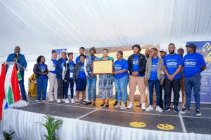 A group of participants wearing matching blue “Future Skills Competition” T-shirts stand on a stage during an award presentation. Several participants and event representatives hold a framed certificate, while a speaker stands at a podium decorated with South African flag colors. Event banners and a large display screen are visible in the background.