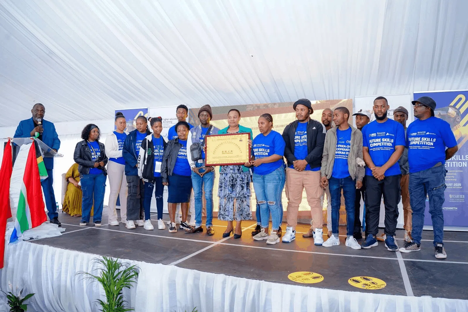 A group of participants wearing matching blue “Future Skills Competition” T-shirts stand on a stage during an award presentation. Several participants and event representatives hold a framed certificate, while a speaker stands at a podium decorated with South African flag colors. Event banners and a large display screen are visible in the background.