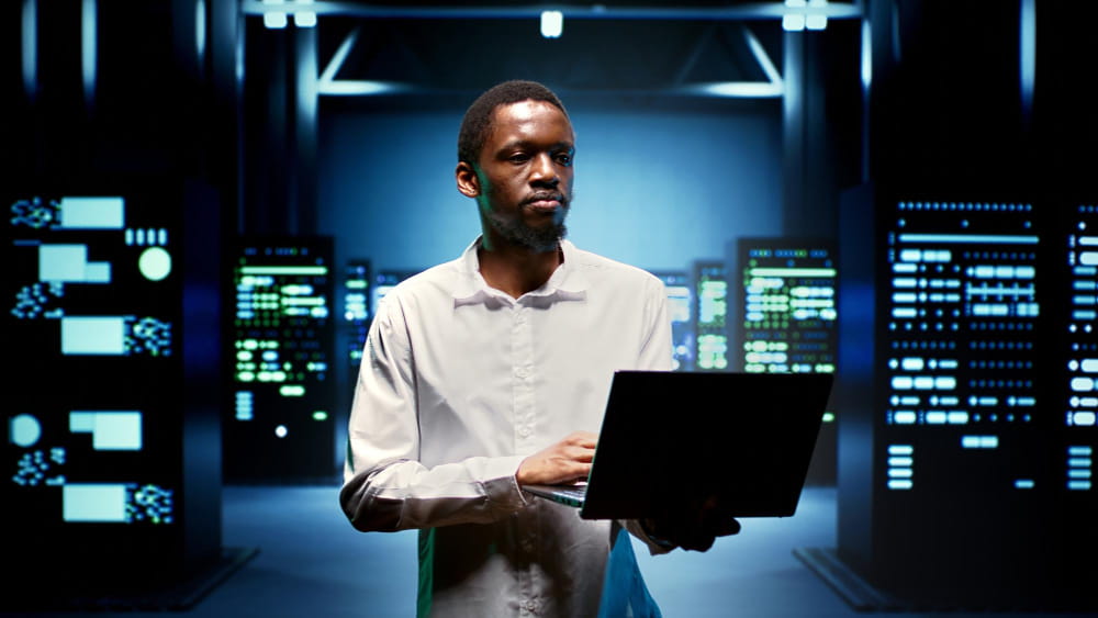 “Person using a laptop in a blue-lit server room surrounded by computer equipment and illuminated server racks.”