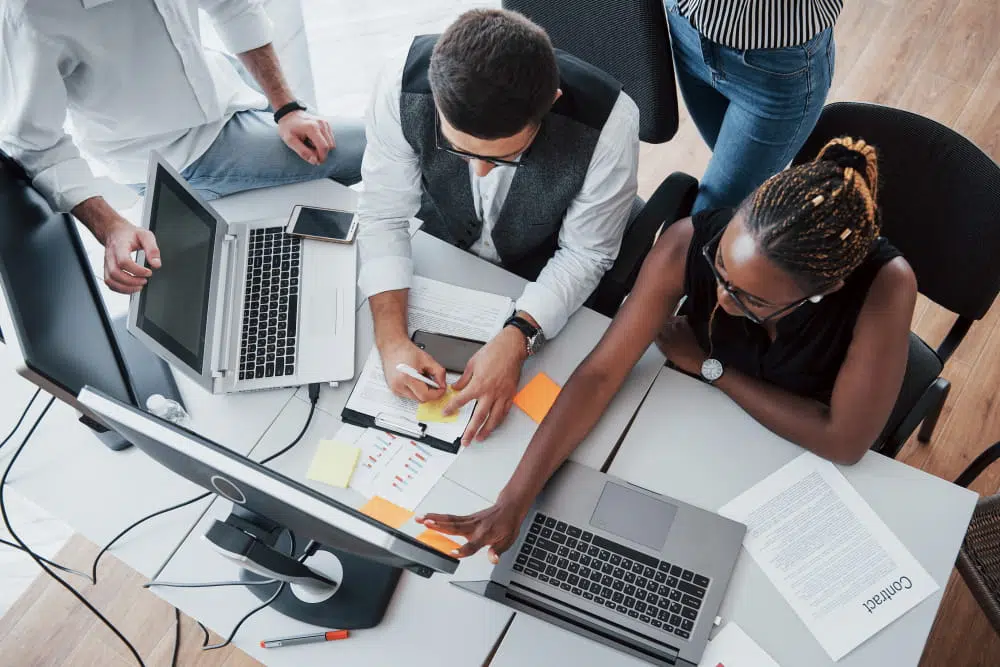 Overhead view of a collaborative office workspace where four people are gathered around a desk with laptops, computer monitors, documents, sticky notes, and a smartphone. One person is writing on a sticky note while another points towards materials on the desk, suggesting a team discussion or project planning session. A printed document labelled “Contract” is visible on the table.