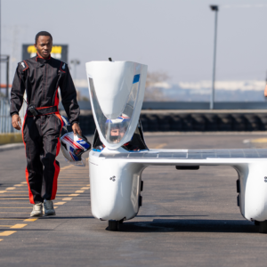 A person in a black and red racing suit walks along a racetrack holding a helmet beside a white, streamlined solar-powered vehicle. Another helmeted occupant is visible inside the vehicle’s cockpit. The vehicle is parked on a paved track with safety barriers and light poles in the background under a clear sky.