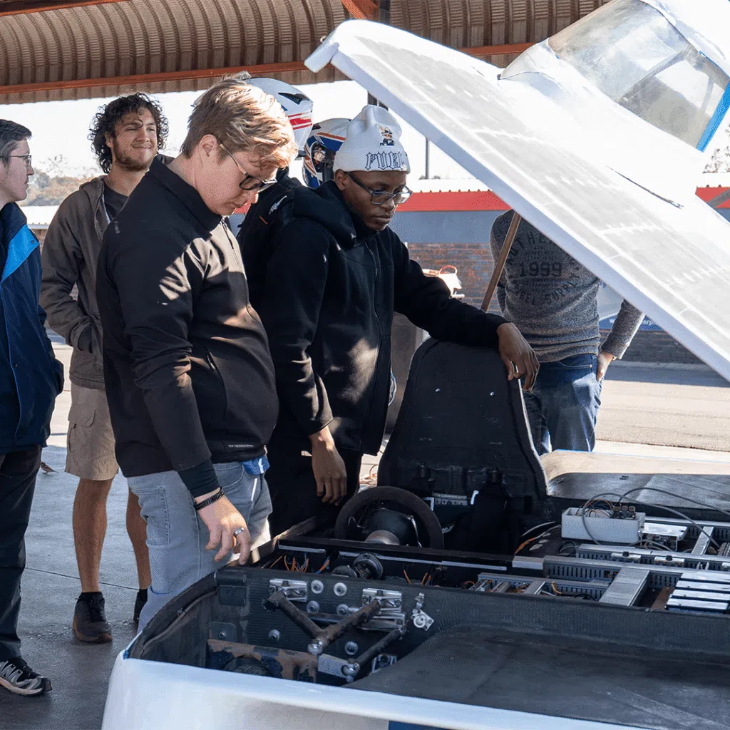 Several people gather around an open solar-electric vehicle under a covered structure. The vehicle’s top panel is raised, revealing the cockpit, seat and internal mechanical and electrical components. Two people stand closest to the exposed interior, examining the vehicle, while others look on from around the car. The vehicle has a lightweight composite body and a solar-panel-covered surface, with bright daylight illuminating the scene.