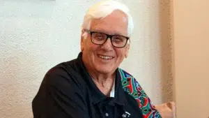 Portrait of a person with short white hair and black-framed glasses, seated indoors against a light textured wall. The person is wearing a dark collared shirt with a colorful patterned panel on one shoulder and is facing the camera. Soft indoor lighting illuminates the scene, and part of a light-colored cushion is visible in the background.