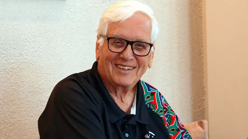 Portrait of a person with short white hair and black-framed glasses, seated indoors against a light textured wall. The person is wearing a dark collared shirt with a colorful patterned panel on one shoulder and is facing the camera. Soft indoor lighting illuminates the scene, and part of a light-colored cushion is visible in the background.