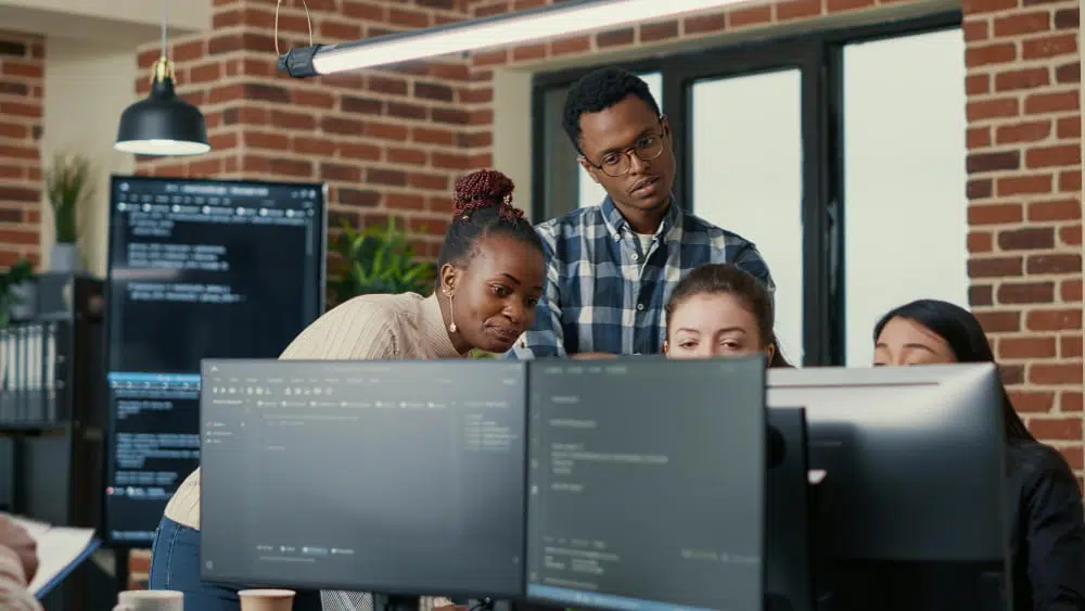A group of people working together in a modern office environment, gathered around multiple computer monitors displaying software development tools and code. Additional screens in the background show programming interfaces, while the workspace features exposed brick walls, large windows, and collaborative technology-focused workstations.