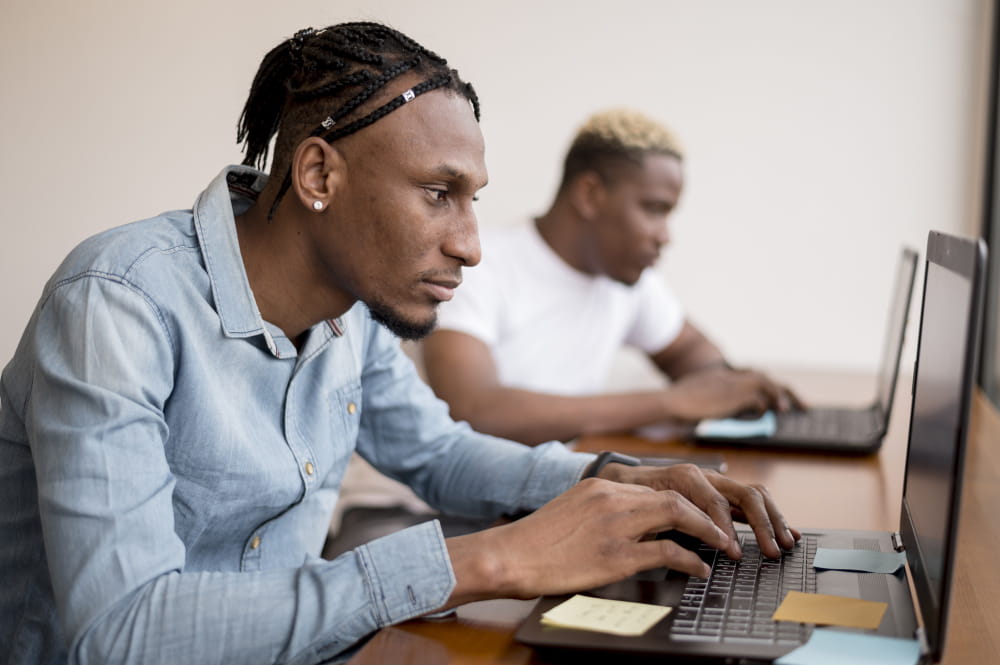 Two people working on laptops at a desk, with one person in the foreground typing and another working in the background