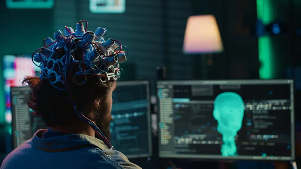 Researcher using a brain-computer interface headset to analyse machine learning and neural data on computer screens in an AI laboratory.