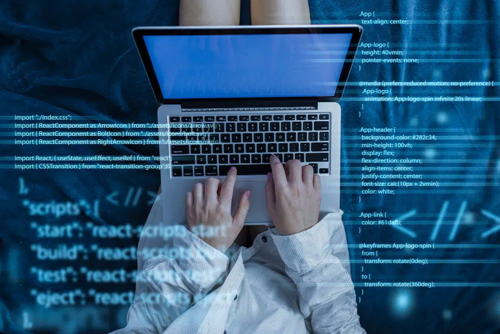 Person typing on a laptop viewed from above, with blue computer programming code and software scripts overlaid across the image